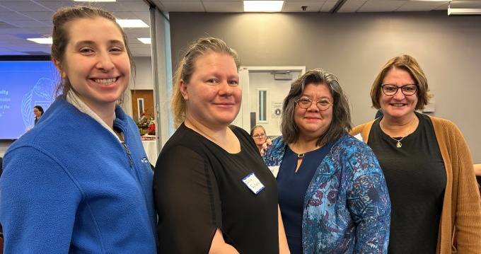 Four adults stand together indoors, smiling and conversing, wearing business casual attire in a hallway or event space.