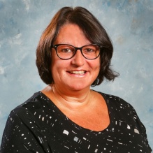Studio portrait of an adult wearing a black patterned short sleeve top, photographed against a mottled light blue background.