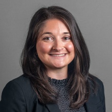 Portrait of Stacy Walker wearing dark grey jacket, black and white polkadot top, with a grey backdrop.