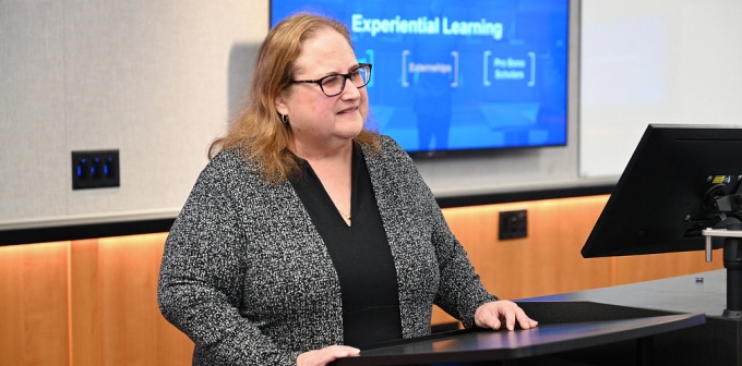 Person standing at a lectern in a classroom or conference room, speaking while a presentation slide is displayed on a screen behind them. The slide includes the title “Experiential Learning” with additional text beneath it.