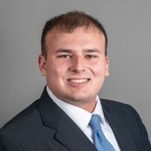 Close‑cropped professional headshot of a person wearing a dark suit jacket, white dress shirt, and light blue tie, photographed against a plain gray background.