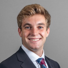 Close‑cropped professional headshot of a person wearing a dark suit jacket, white dress shirt, and patterned tie, photographed against a plain gray background.
