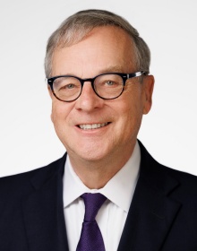 A person wearing a dark suit jacket, white dress shirt, and a solid purple necktie, posed against a plain light gray background.