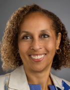 A person with short, curly hair wearing a light-colored blazer with blue accents over a blue textured top, posed against a plain gray background.