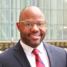 Man standing outside, smiling, wearing a suit and tie. 