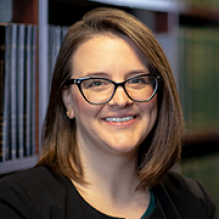 A person with shoulder‑length brown hair wearing a dark blazer sits indoors in front of bookshelves filled with books.