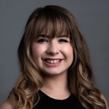A person with shoulder length wavy light brown hair wearing a sleeveless black top, posed against a plain gray background.