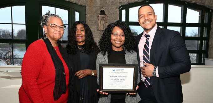Four people stand together indoors in front of large green‑framed windows. The person second from the right holds a framed certificate, while the others stand beside them in a group pose. The setting appears to be an awards or recognition event.