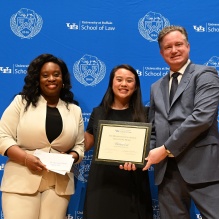 Three people stand in front of a University at Buffalo School of Law backdrop. The person in the center holds a framed certificate while the person on the left holds an envelope. The person on the right stands beside them in a suit. The group appears to be participating in an awards or recognition event.