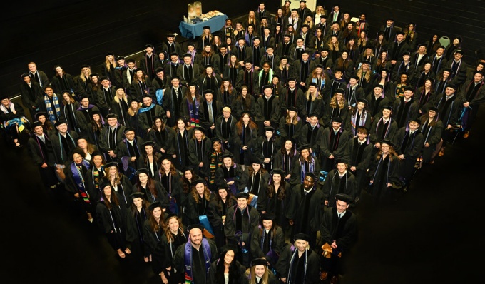 A room full of future graduates standing in several rows, image taken from above looking down of young adults wearing graduation regalia, look up, smiling at the camera. 