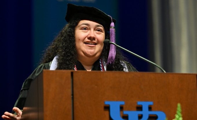 A commencement speaker in academic regalia stands at a podium with the University at Buffalo logo, delivering remarks. A UB banner hangs in the background, and floral arrangements and a ceremonial mace are visible in front of the podium.