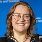 Sara Bachraty wearing glasses, black and white jacket, standing in front of a blue background.